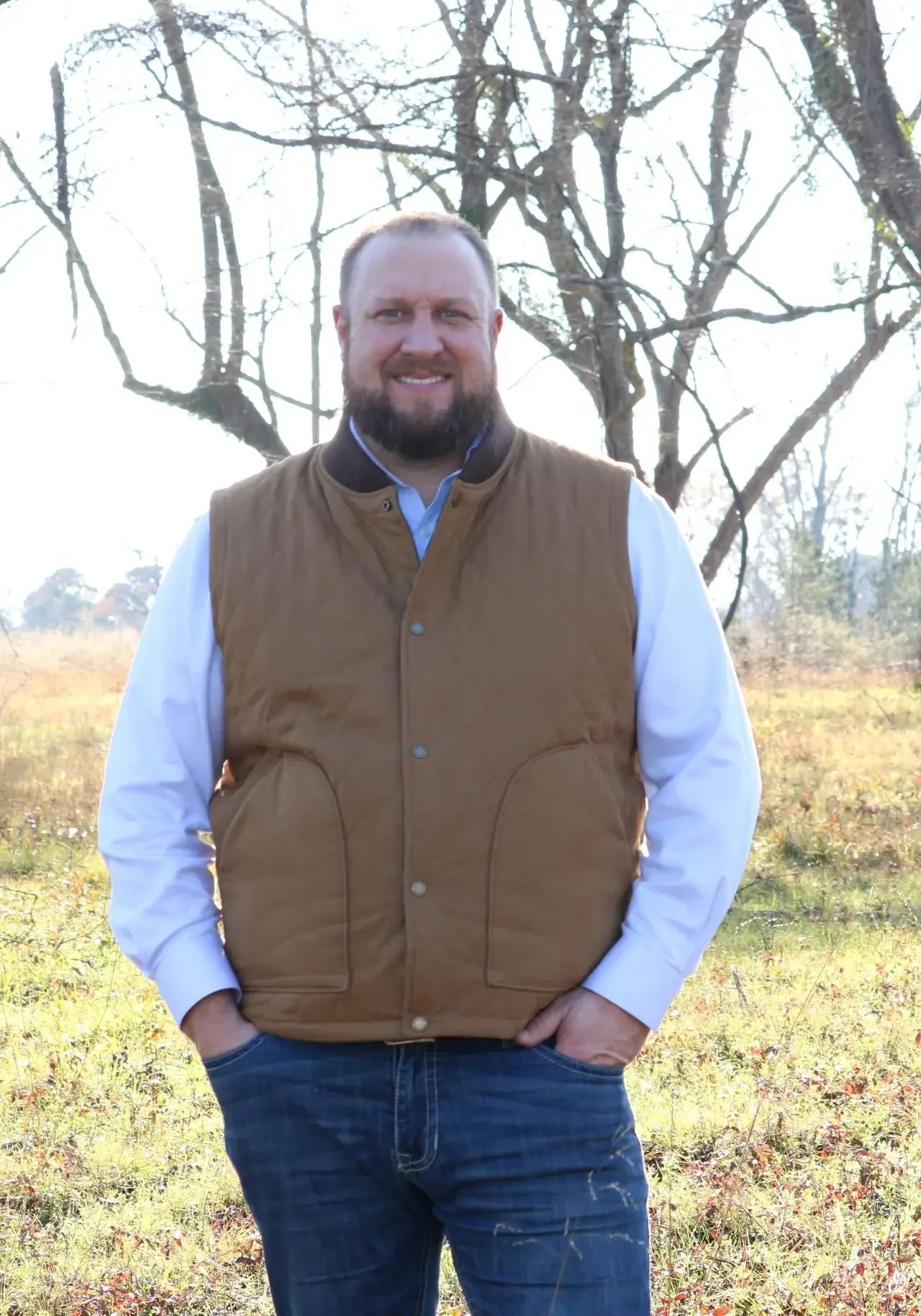 Man in a brown vest standing outdoors in a sunny, leafless tree area.